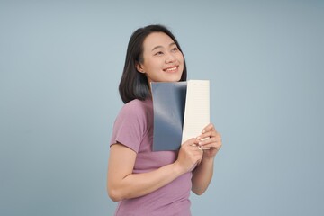 Naklejka premium Smiling Asian woman holds notebooks while standing against a light blue background in a casual setting