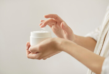 Closeup of young woman touching cream with finger isolated on white background. Female person holding jar with moisturizing white cream in hands. Concept of body health and skin care.