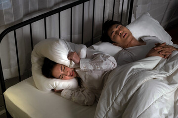 Woman struggles to sleep beside snoring partner, covering ears with pillow in dark bedroom. Concept...
