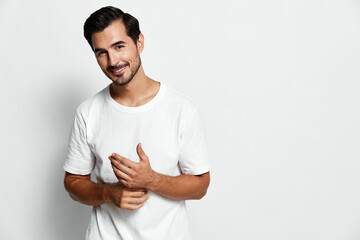 Smiling young man in casual white t-shirt stands against a plain light background, looking at the camera with a friendly expression. People lifestyle concept.