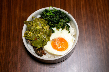 A bowl of rice with side dishes, likely dhal, green vegetables, and a boiled egg, arranged on a wooden table, creating a simple yet appetizing meal