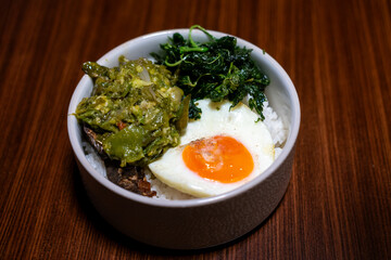 A bowl of rice with side dishes, likely dhal, green vegetables, and a boiled egg, arranged on a wooden table, creating a simple yet appetizing meal