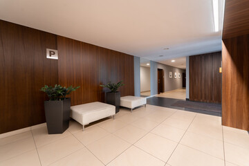 Hallway of a modern residential building with wood panels and tile floor. This spacious corridor features a seating area with ottomans and plants. Clean lines mark its contemporary interior design.