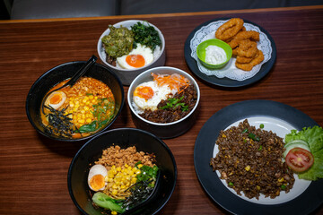 A variety of Japanese dishes, possibly from a restaurant menu, featuring ramen, rice bowls with egg and toppings, and fried appetizers, arranged on a wooden table.