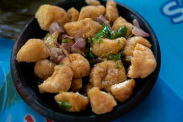 a serving of Tahu Gejrot, a popular Indonesian street food,  the fried tofu pieces mixed with a dark, watery sauce, slices of shallots, and green chili peppers in a dark bowl
