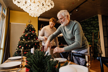 A senior White man and his adult White son preparing a festive dining table for Christmas dinner in a warmly lit home next to a decorated Christmas tree with red ornaments and string lights.