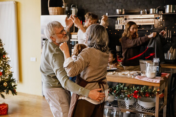 An elderly white man and woman dance together in a decorated kitchen with a Christmas tree. The family members, including women and children, prepare food and celebrate the holiday indoors.