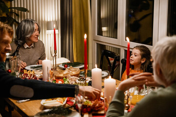 An elderly White woman, an elderly White man, an adult White man, and a school-aged girl sit around a festive dinner table indoors, celebrating Christmas with food, candles, and holiday decorations.