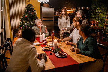 A multinational group of people of different ages gathered around a decorated wooden dining table, enjoying a celebration in a cozy home interior with candles, food, and a lit Christmas tree.