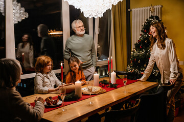 A big White family of multiple generations gathered at a festively decorated Christmas dinner table in the evening, smiling and talking near a lit Christmas tree and candles in a modern dining room.