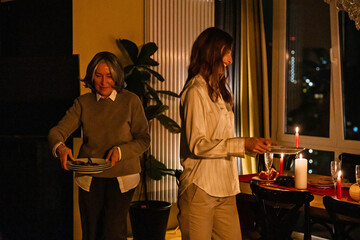 A caucasian woman and a young woman stand in a modern apartment kitchen at night, placing plates on a festive dinner table with candles and drinks during a Christmas celebration.