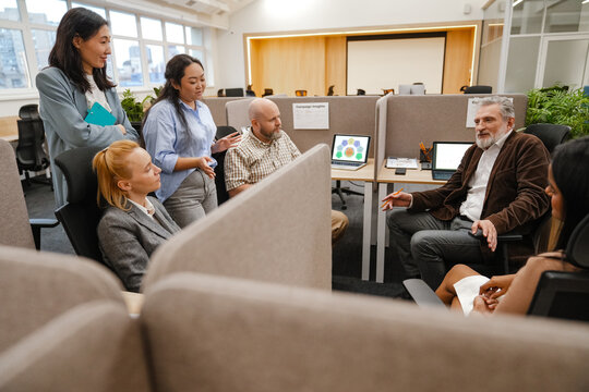 A multiracial group of professionals, including a middle-aged Caucasian man and diverse team members, sit and stand in a modern office cubicle area during a collaborative meeting. - Powered by Adobe