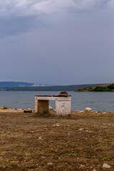 An old bench with a hat on top stands alone by a lake under a cloudy sky, surrounded by dry ground...