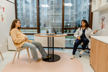 An Indian female doctor is sitting on a chair and pointing with a stick at a poster of a pregnant woman on a wall while looking at a White pregnant woman who is sitting at a table in front of her