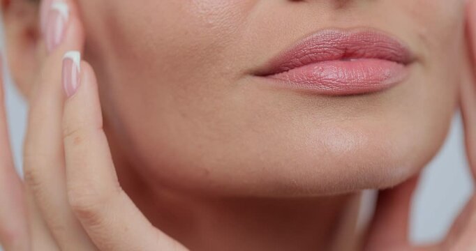Close up feminine lips and chin applying anti aging cream on face skin. Women demonstrate moisturizing facial care touching cheek isolated on gray studio background. Skincare concept.