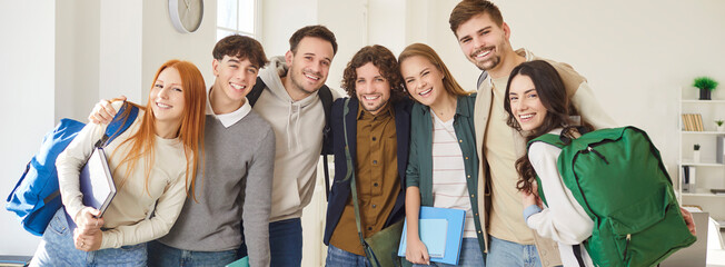 Obraz premium Group portrait of happy smiling students standing together in classroom and hugging each other. Cheerful young people carrying backpacks and notebooks, looking at camera, ready for learning. Banner.
