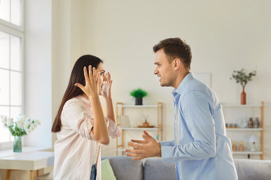Young angry couple man and woman standing at home and arguing in quarrel. Unhappy husband and wife quarreling in the living room. Marriage problems, relationship and divorce concept. - Powered by Adobe
