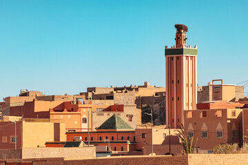 Traditional Moroccan architecture featuring a prominent minaret rising above the rooftops of a town, bathed in the warm sunlight of a clear blue sky