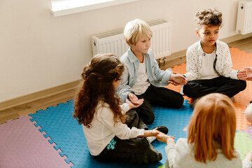 A multinational group of pupils sitting cross-legged on colorful foam mats in a classroom, holding hands in a circle, engaging in a mindfulness or relaxation exercise while wearing casual clothes.