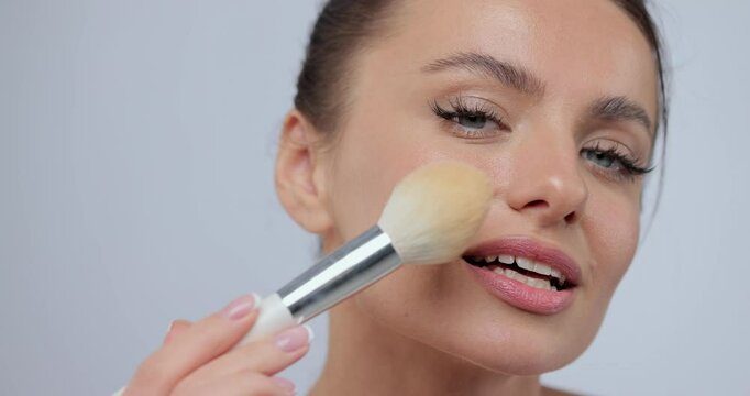 Young woman applying makeup blush with brush across her face while standing against studio gray background. Concept of cosmetology, skincare.