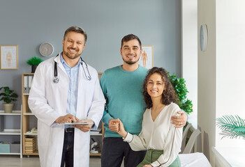 Obraz premium Portrait of smiling happy young couple husband and wife standing with a male family doctor therapist in medical clinic or hospital. Health care, family medicine and check up concept.