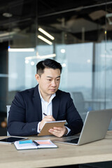 Fototapeta premium Asian businessman in suit working at his desk in the office environment, taking notes.