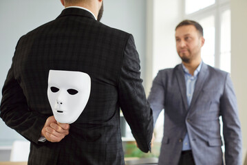 Back view of man in suit hiding white mask behind his back while shaking hands with male colleague...