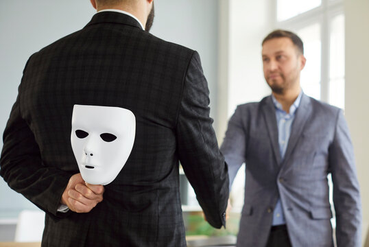 Back view of man in suit hiding white mask behind his back while shaking hands with male colleague in office. Concept of hypocrisy, deception, dishonesty, false promises or insincerity in partnership.