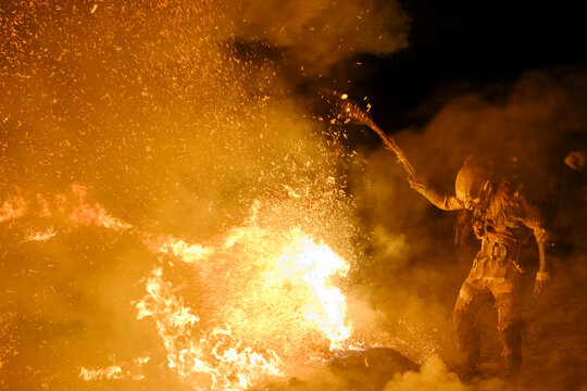 A Krampus figure stands near a large fire, holding a torch aloft, surrounded by sparks and smoke. Krampus and St Nicholas, 5 December in Tarvisio, Friuli Venezia Giulia, Julian Alps, Italy.