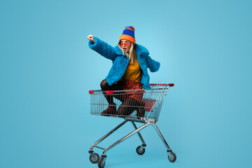 Side view of crazy trendy young female in bright clothes holding paper bags and riding shopping...