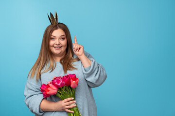 Charming plump female with bouquet of beautiful tulips and paper crown smiling and looking at camera while pointing up on blue background