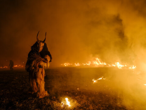 A Krampus figure in a furry suit and horned mask stands amidst smoke and fire. Another figure is visible in the background. Krampus and St Nicholas, 5 December in Tarvisio, Italy.