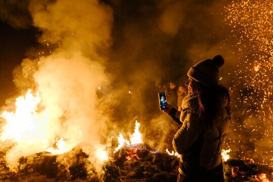 A woman in a winter hat and coat takes a photo with her phone of a large bonfire. Sparks fly in the air. Krampus and St Nicholas, 5 December in Tarvisio, Friuli Venezia Giulia, Julian Alps, Italy.