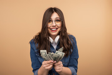 A joyful young woman with glasses, smiling and holding a handful of cash against a beige background, conveying happiness, financial luck, and a positive attitude.