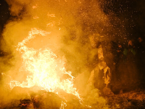 A large bonfire with bright flames and smoke fills the frame. A figure in a Krampus mask is visible in the smoke. Krampus and St Nicholas, 5 December in Tarvisio, 