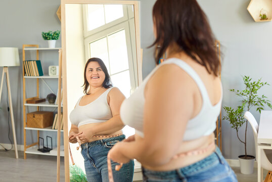 Confident plus size woman measuring waist with tape measure while looking at reflection in mirror at home. Smiling overweight female checking weight loss progress, feeling empowered and motivated.