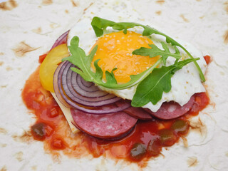 Unwrapped thin pita bread with fried egg with arugula leaf on slices of sausage, cheese, rings of red onion and yellow paprika for grill. Variant of healthy summer meal and hearty breakfast.