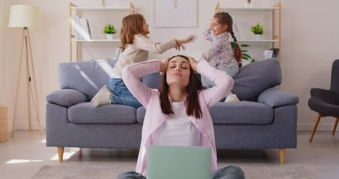 Tired and stressed mother attempts job work on laptop at home while kids misbehave with noise, creating chaos in a challenging environment with focus on balancing responsibilities and stress.