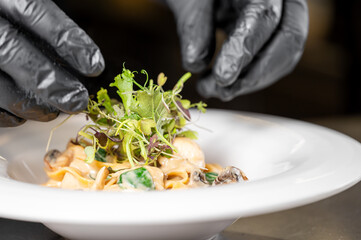 A chef's hands in black gloves carefully place fresh microgreens on a gourmet pasta dish in a white bowl, showcasing culinary artistry and meticulous plating in fine dining.