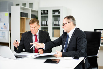 Businessmen in formal suits planning project strategy with charts, laptop, and tablet in modern office. Collaboration, consulting