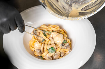 A chef's hand in a black glove plates creamy pasta with mushrooms and spinach using tweezers. Sauce is poured from a pan into a white bowl. Professional culinary preparation in a restaurant kitchen
