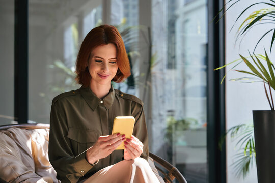 Smiling young professional business woman, happy lady with red hair holding mobile cell phone in hands working sitting in chair in sunny office looking at cellphone and using smartphone.
