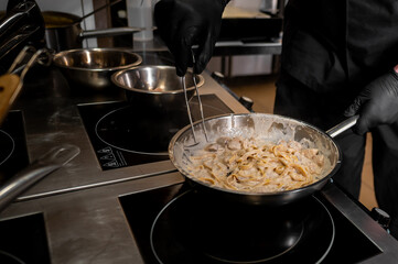 A professional chef in black gloves prepares creamy chicken pasta in a stainless steel pan on an induction stove, surrounded by kitchen tools in a restaurant setting.