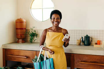 Plakat Brazilian woman smiling and holding a vegetable bag with mobile phone in kitchen at home