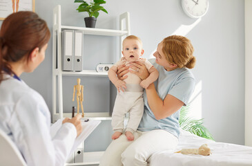 Young smiling mother sitting on couch in medical office, holding toddler baby in her hands during...