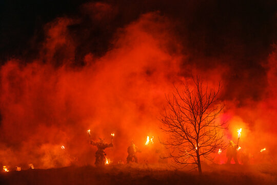 Figures in costumes called Krampus hold torches in a smoky, red-lit landscape. A bare tree stands to the right. Krampus and St Nicholas, 5 December in Camporosso Valcanale, Tarvisio, Italy.