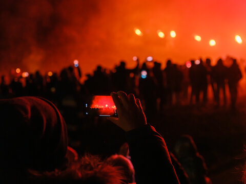 A person holds up a phone to photograph a crowd of people silhouetted against a fiery red sky. Krampus and St Nicholas, 5 December in Camporosso Valcanale, Tarvisio, Italy.