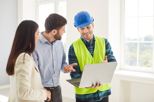 Happy couple husband and wife homeowners together with foreman, construction engineer, architect standing indoors, looking at digital house remodeling plan on laptop, discussing interior redesign