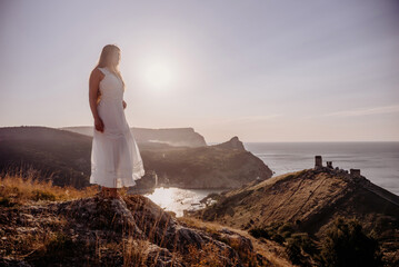A woman stands on a rocky hill overlooking the ocean. She is wearing a white dress and she is enjoying the view. The scene is serene and peaceful, with the sun shining brightly in the background.