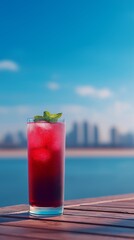 Obraz premium Berry Refresher by the Sea: A vibrant red cocktail garnished with a sprig of mint, sits refreshingly on a wooden table against a backdrop of sparkling blue water and a distant cityscape.